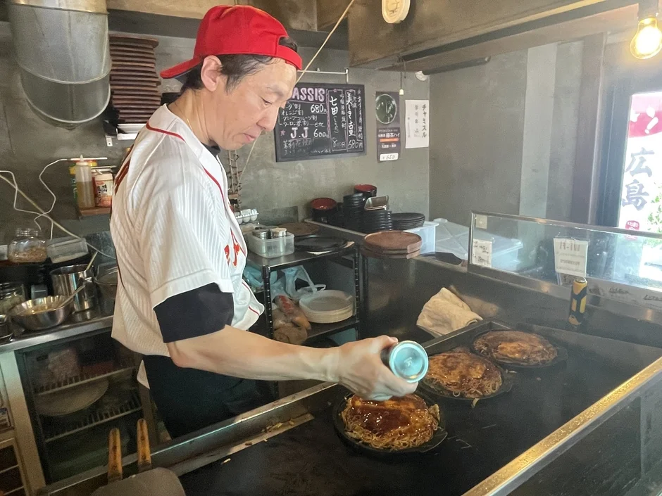 赤い帽子と野球のユニフォームのようなシャツを着た男性が、鉄板の上で複数のお好み焼きにソースをかけて調理している。