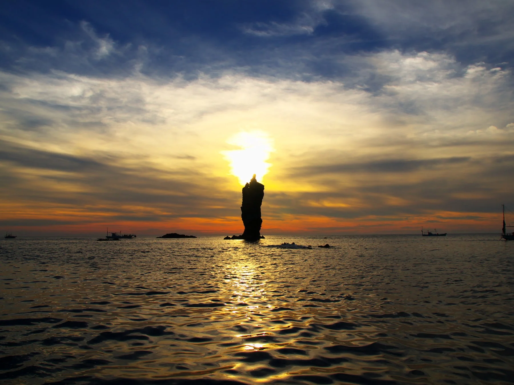 夕暮れ時の海にそびえ立つ奇岩のシルエットが印象的な一枚。燃えるような夕日が岩の背後で輝き、空と水面をオレンジ色に染めている。遠くには数隻の船が見え、穏やかな海の広がりを感じさせる美しい風景。