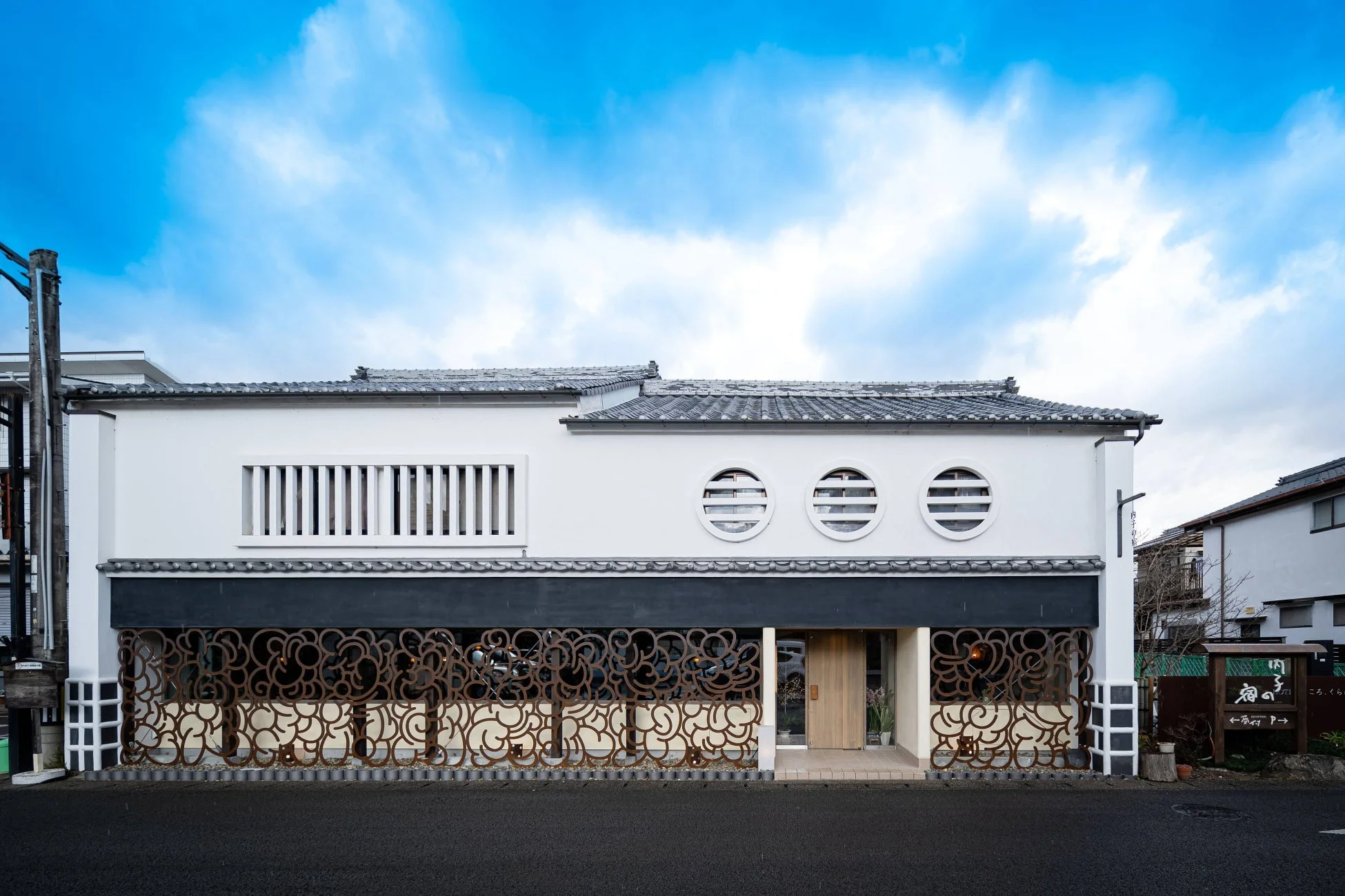 白い壁と瓦屋根が特徴的な、日本の伝統とモダンが融合した建築物。装飾的な金属製スクリーンが目を引く店舗のファサードが、青い空の下にたたずむ様子。