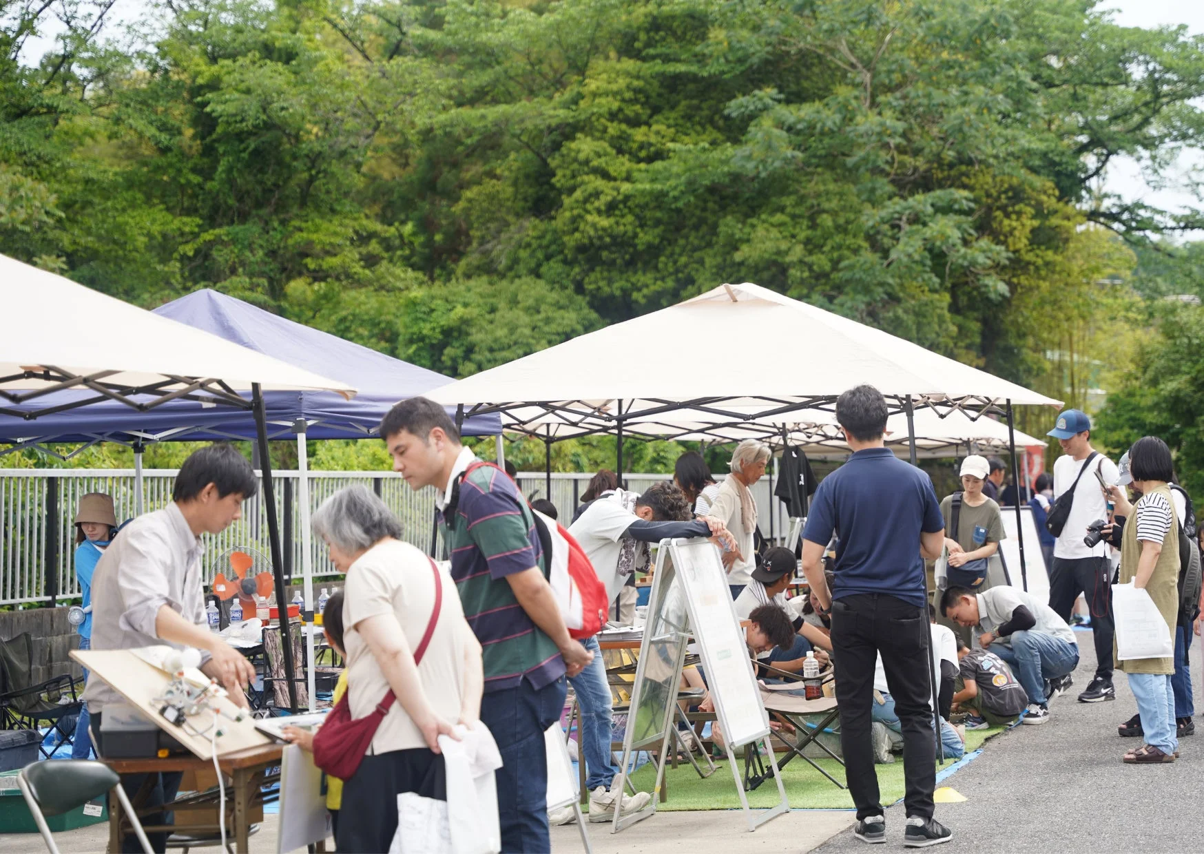 緑豊かな木々を背景に、多くの人々がテントの下に集まっている屋外イベントの様子