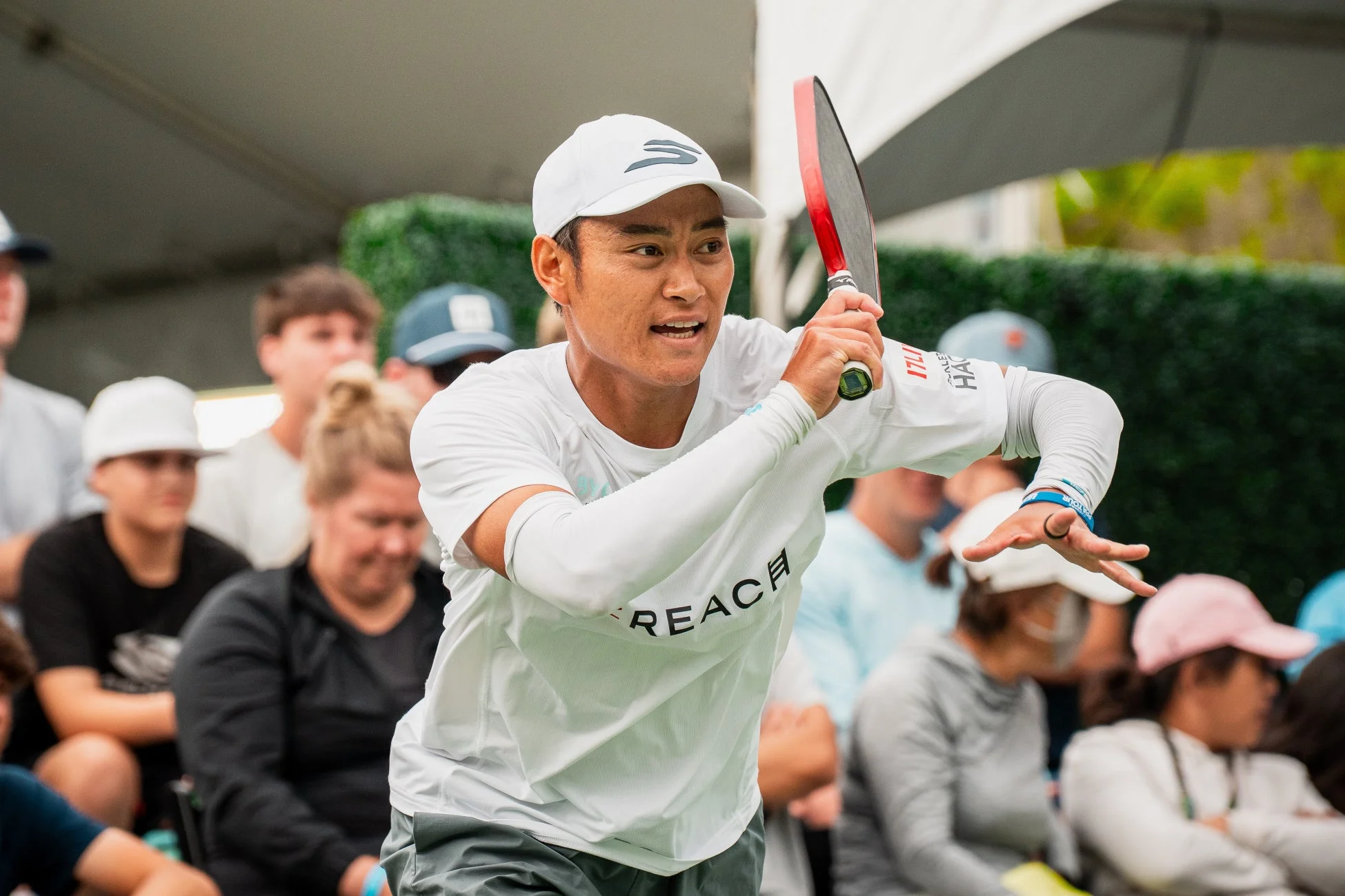 白い帽子と白いスポーツシャツを着たアジア系の男性選手が、赤いピックルボールのラケットを構え、試合中に集中している様子です。