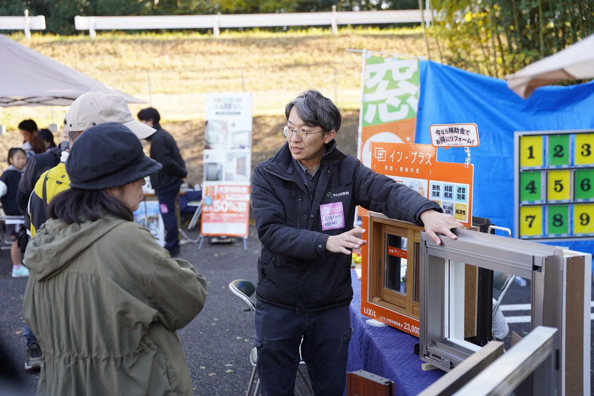 屋外のイベント会場で、リフォーム会社の担当者が顧客にLIXILの窓「インプラス」について説明している様子