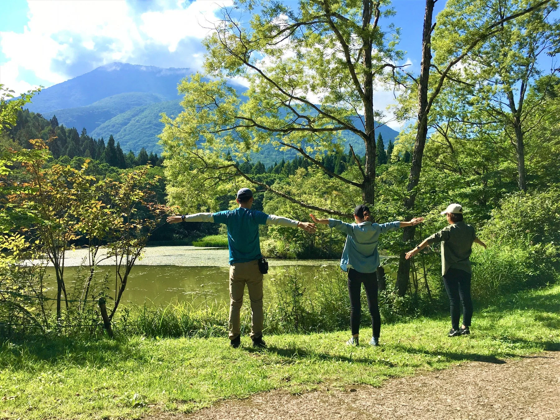 湖と山を背景に、3人の人物が両腕を広げて自然を満喫している様子。青空の下、豊かな緑に囲まれた開放的な風景です。