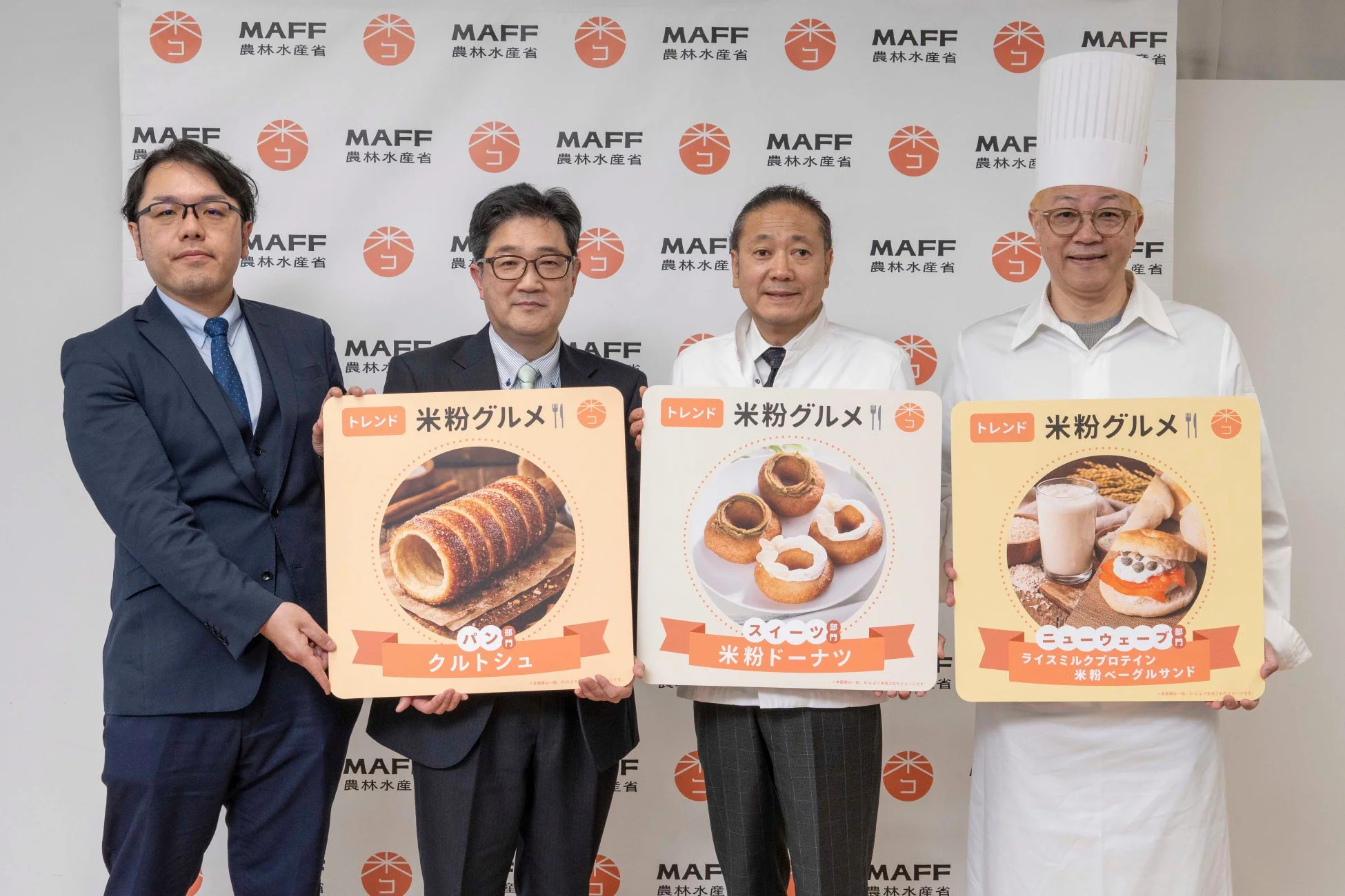 農林水産省のロゴを背景に、4人の男性が「米粉グルメ」の新製品を紹介している集合写真です。パン、スイーツ、ニューウェーブの3部門で、クルトシュ、米粉ドーナツ、ライスミルクプロテイン米粉ベーグルサンドが展示されています。