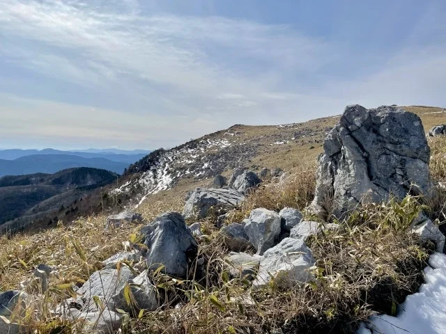 岩が点在する山頂付近の広大な景色です。枯れ草が生い茂り、地面の一部には雪が見られます。遠くには連なる山々が霞んで見え、空には白い雲が浮かんでいます。