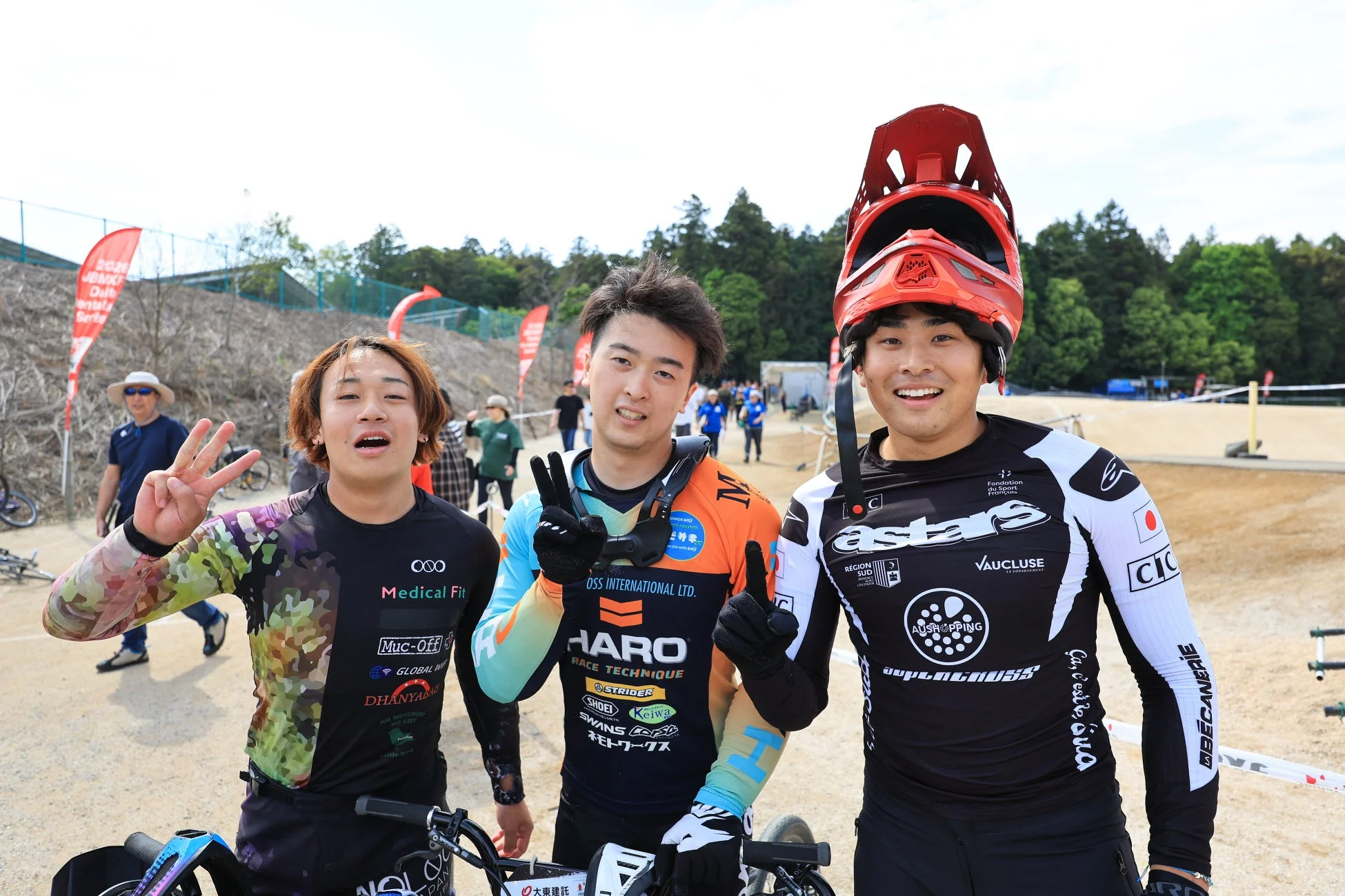 BMXレース会場で、3人の若いBMXライダーが笑顔でポーズをとっています。彼らはそれぞれのスポンサーロゴが入ったレースウェアを着用しており、活気ある大会の雰囲気が伝わってきます。