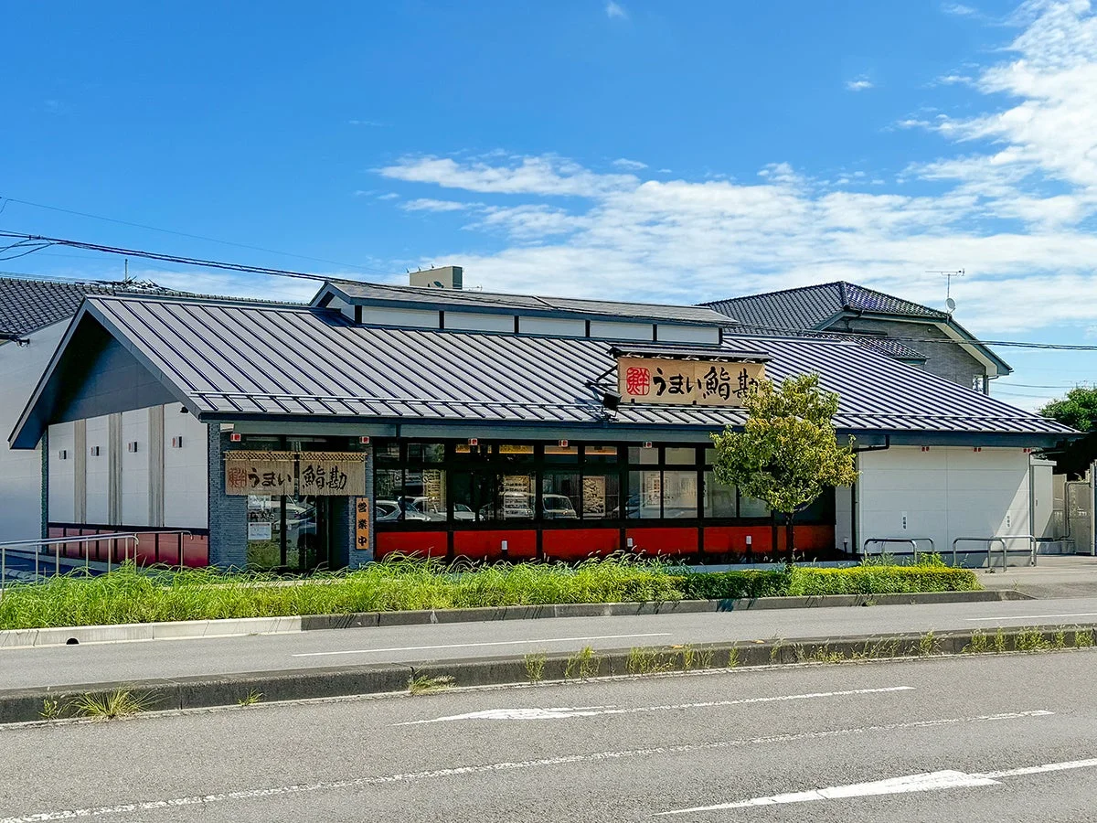 「うまい鰻勘」という看板のある鰻料理店の外観を捉えた写真です。モダンな建物で、手前には道路と植え込みがあり、晴れた青空が広がっています。