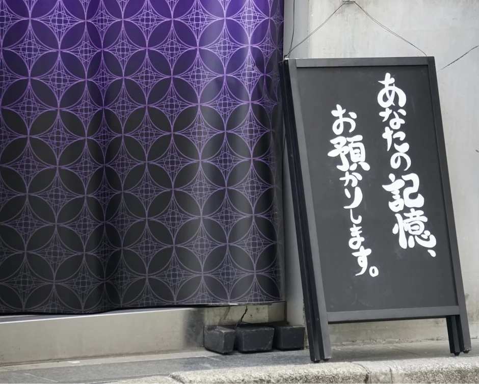 伝統的な和柄の紫色の暖簾と「あなたの記憶、お預かりします。」と書かれた黒いA型看板
