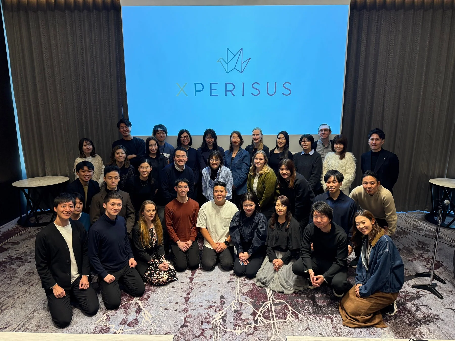 XPERISUSのロゴが映るスクリーンを背景に、多くのメンバーが笑顔でポーズをとっている集合写真