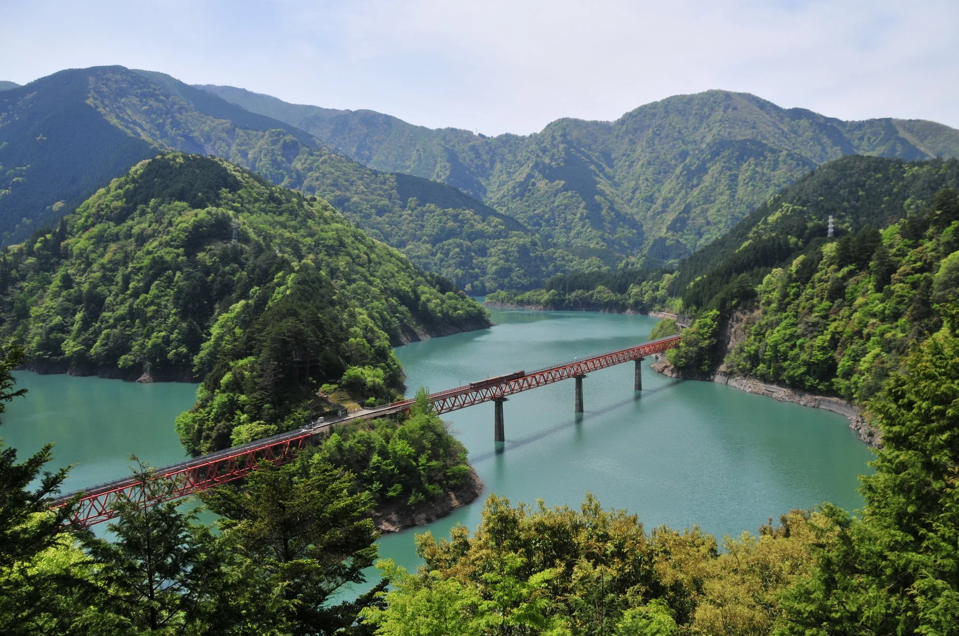 新緑の山々に囲まれたエメラルドグリーンの湖に、赤い鉄道橋が架かる雄大な自然景観です。静かで美しい日本の風景が広がっています。