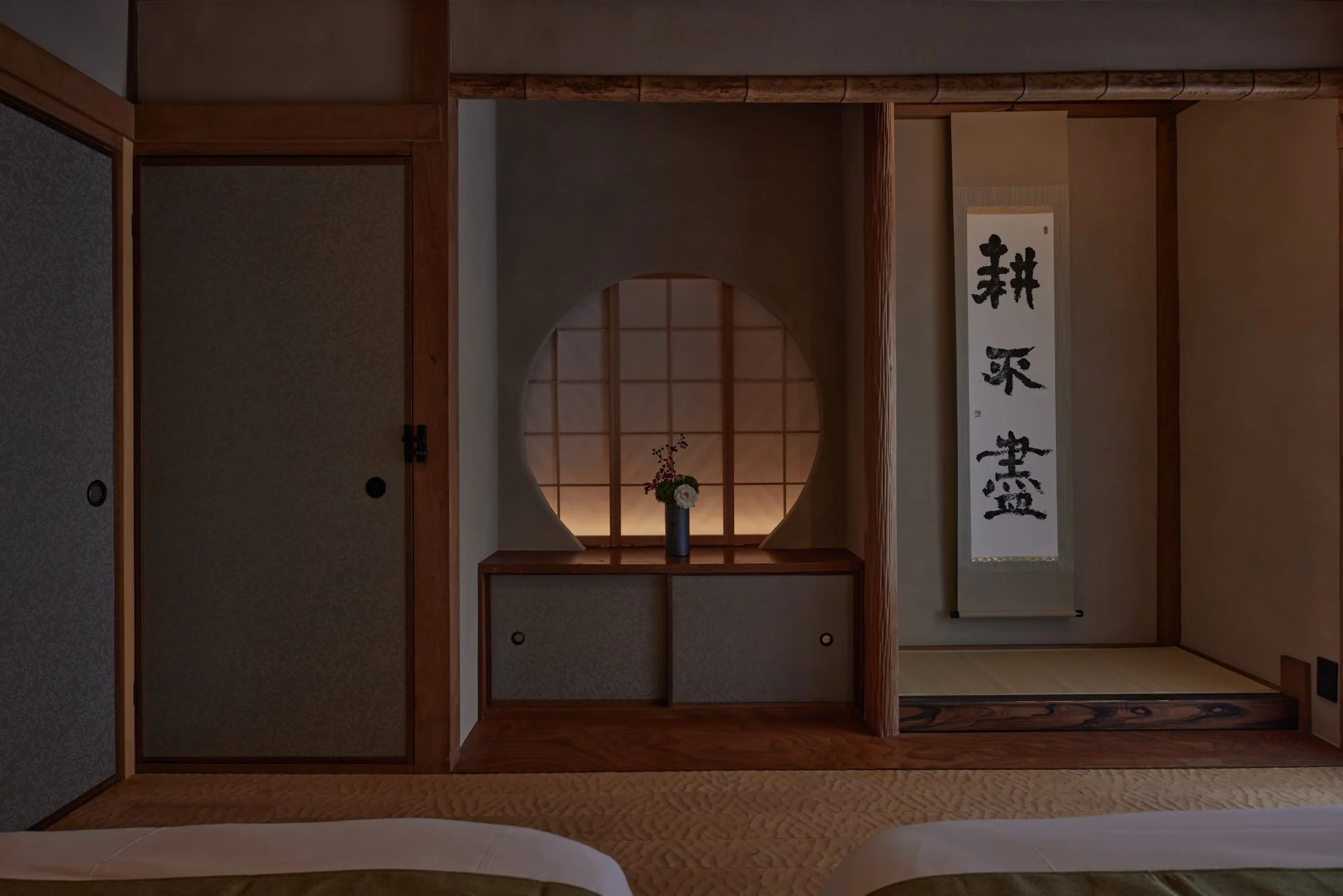 伝統的な和室の落ち着いた空間。床の間には掛け軸と生け花が飾られ、その奥には柔らかな光が漏れる円窓が見える