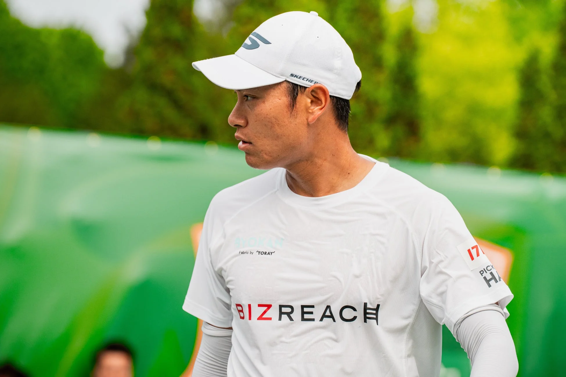 白いキャップとTシャツを着用した男性アスリートが、屋外の緑色のコートで競技中に左方向を見ている。