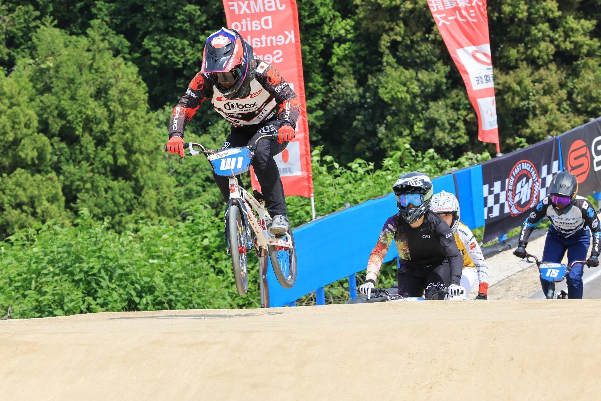 BMXレースで、ライダーがダートトラックのジャンプ台を飛び越えている瞬間を捉えた写真です。複数の選手が競技中で、背景には緑豊かな木々とスポンサーの旗が見えます。