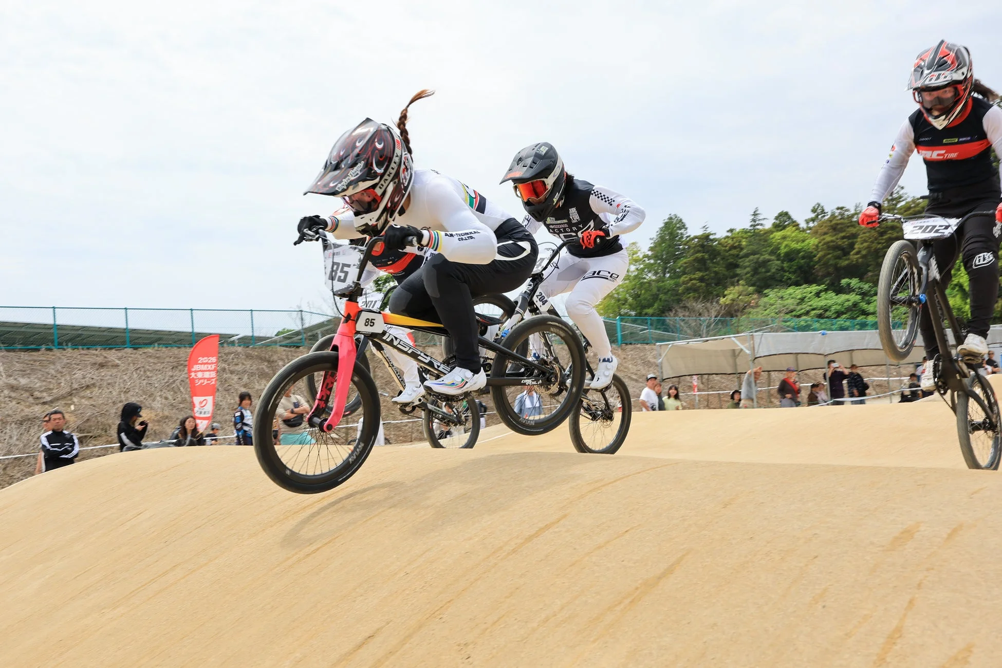 BMXレースで選手たちがダートコースのコブをジャンプしている瞬間を捉えた迫力ある写真です。3人の選手がヘルメットとウェアを着用し、真剣な表情で競技に挑んでいます。背景には観客と緑が見えます。