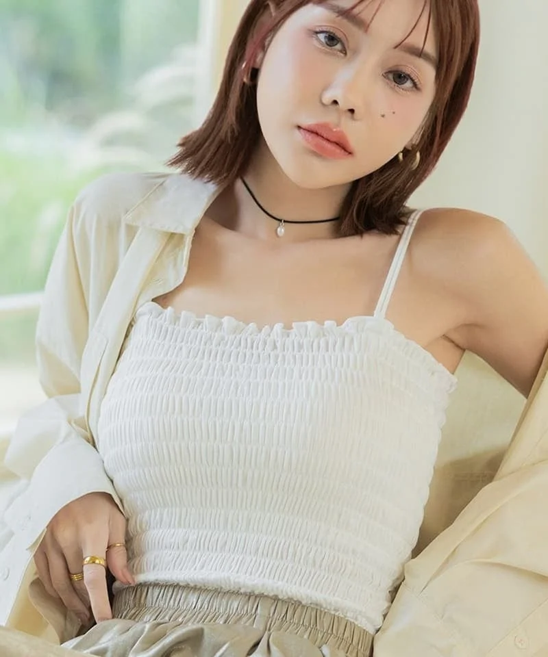 白いシャーリングトップスと淡い色のシャツを着用したショートヘアの女性が、自然光の差し込む屋内でリラックスした表情を見せています。