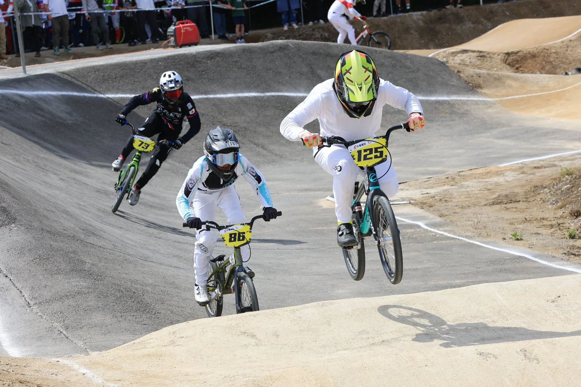BMXレースで3人のライダーがダートコースを走行している様子。手前のライダーはジャンプしており、ダイナミックな瞬間を捉えています。