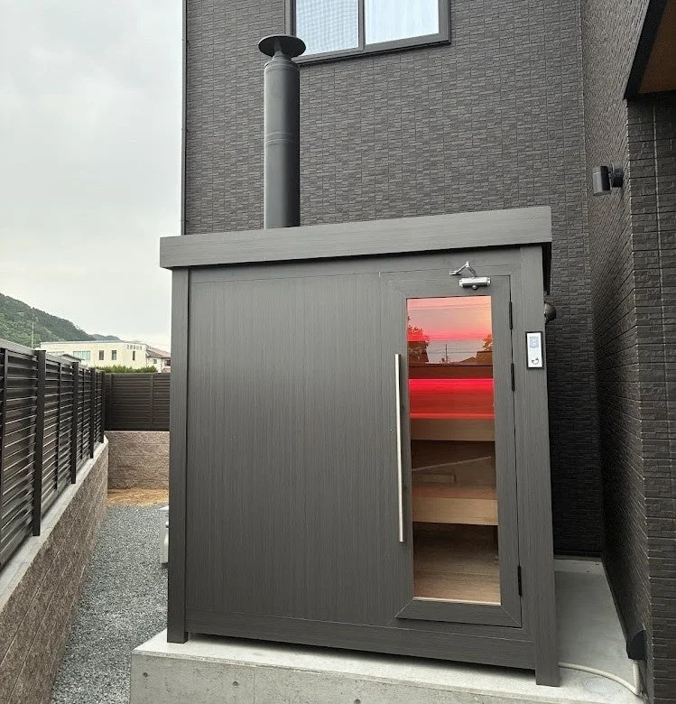 モダンな住宅の隣に設置されたダークカラーの屋外サウナ小屋です。煙突があり、ガラス扉からは赤い室内灯が見えており、使用中の様子が伺えます。