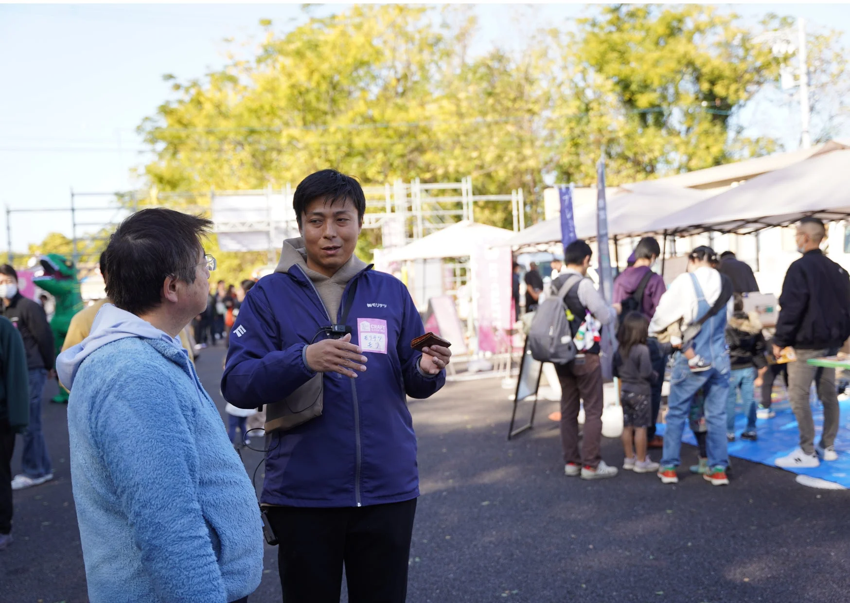 屋外イベントで二人の男性が会話しており、背景には多くの人々やテント、黄葉した木々が見える