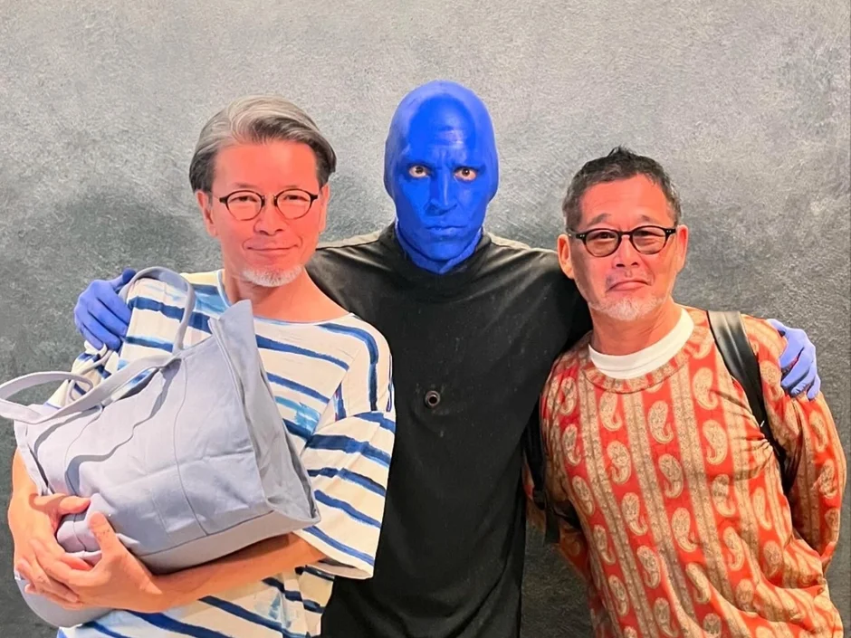 青い顔の男性と2人の男性が肩を組んでポーズを取っている画像。ブルーマンとファンが一緒に記念撮影をしているように見える。