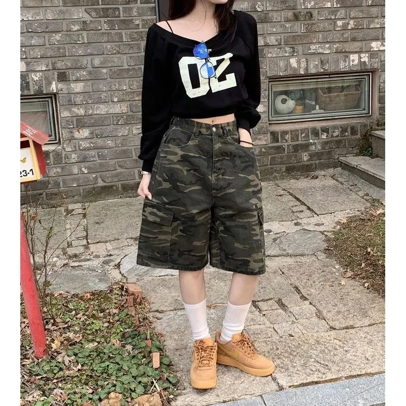女性が黒いオフショルダートップスと迷彩柄のカーゴショーツを着用し、茶色のスニーカーを合わせたカジュアルなストリートファッションで屋外に立っています。