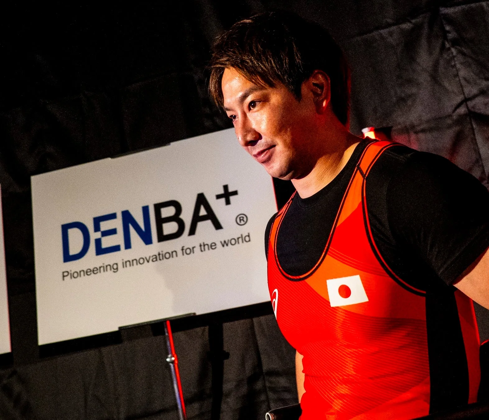 日本の国旗が描かれた赤いユニフォームを着た男性アスリートが、DENBA+のロゴがあるボードの前で微笑んでいる様子。