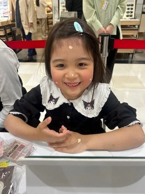 笑顔の幼い女の子が、顔や手にファンデーションらしきクリームを点々とつけ、可愛らしい黒いワンピースを着ている。コスメの試供品を試しているかのような様子で、楽しげな雰囲気が伝わる。