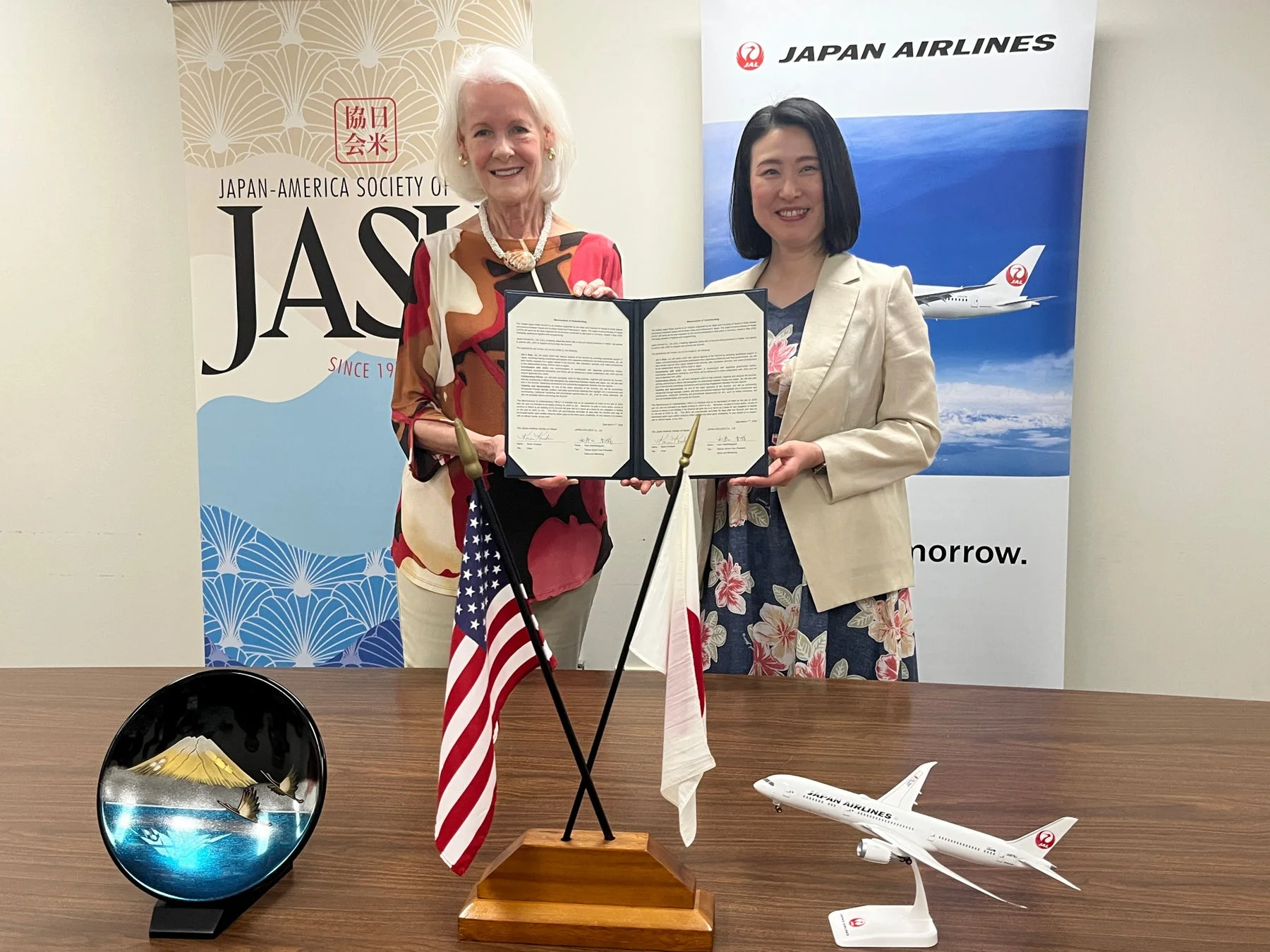 日米協会と日本航空の代表者と思われる2人の女性が、協定書を交わしている様子です。両国の国旗、飛行機の模型、富士山の置物が並べられ、日米間の連携を示すビジネスシーンが描写されています。