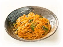 素焼きのような質感の器に盛り付けられた、オレンジ色のソースで和えられたスパゲッティのクローズアップ画像です。具材として緑色の野菜やエビのようなものが見えます。