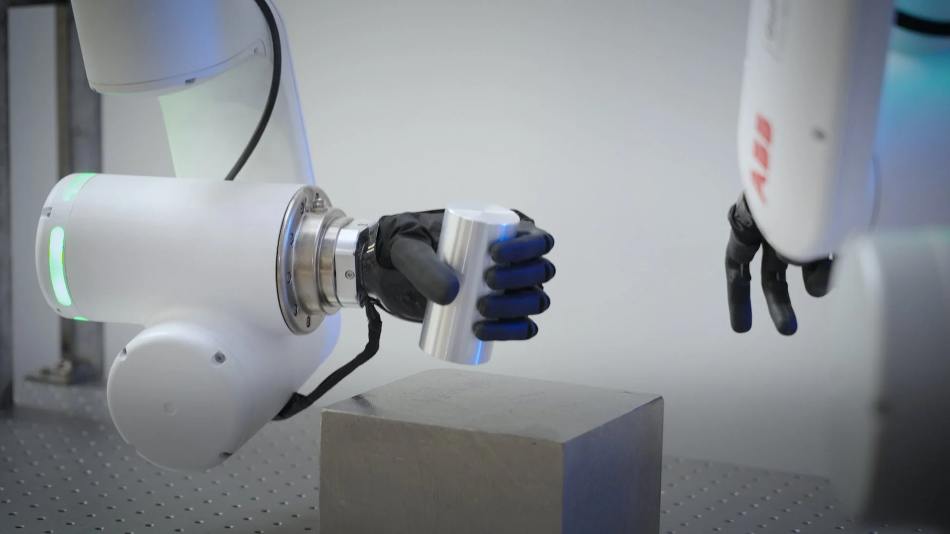 白いロボットアームが黒いグリッパーで銀色の円柱形の物体を掴んでおり、その下には灰色のブロックが置かれている。別のロボットアームも右側に一部見えている。