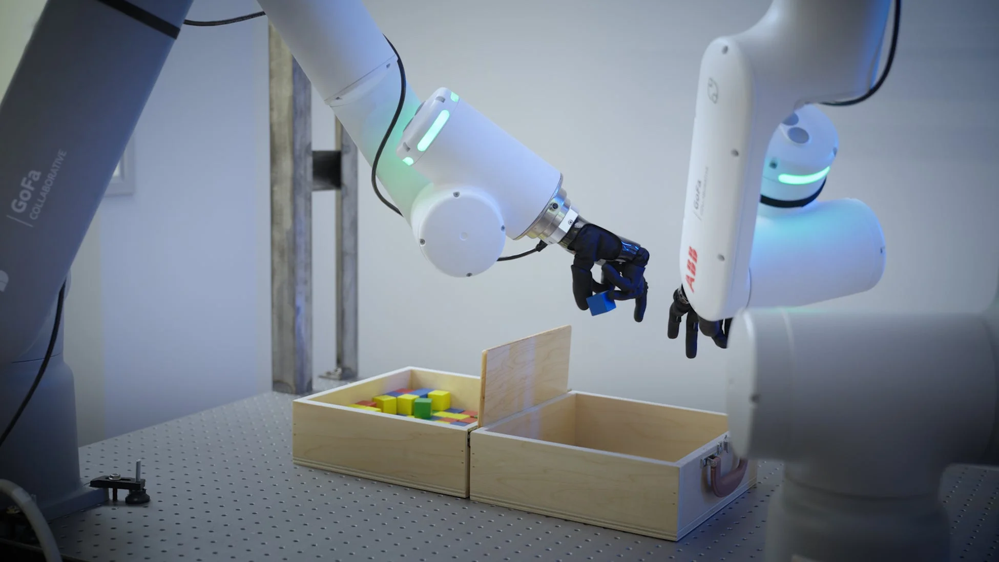画像には、二つの白いロボットアームが写っており、左側のロボットアームが木箱に入ったカラフルなブロックの中から青いブロックを掴もうとしている様子が捉えられています。これは協働ロボットによる精密作業を示唆しており、研究または産業環境での自動化を示しています。