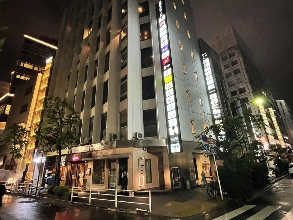 雨上がりの夜、多くの看板が光る都市の街並みが広がる。高層ビルが立ち並び、1階には店舗が見える。路面は濡れており、光が反射している。賑やかな商業地域の夜景である。