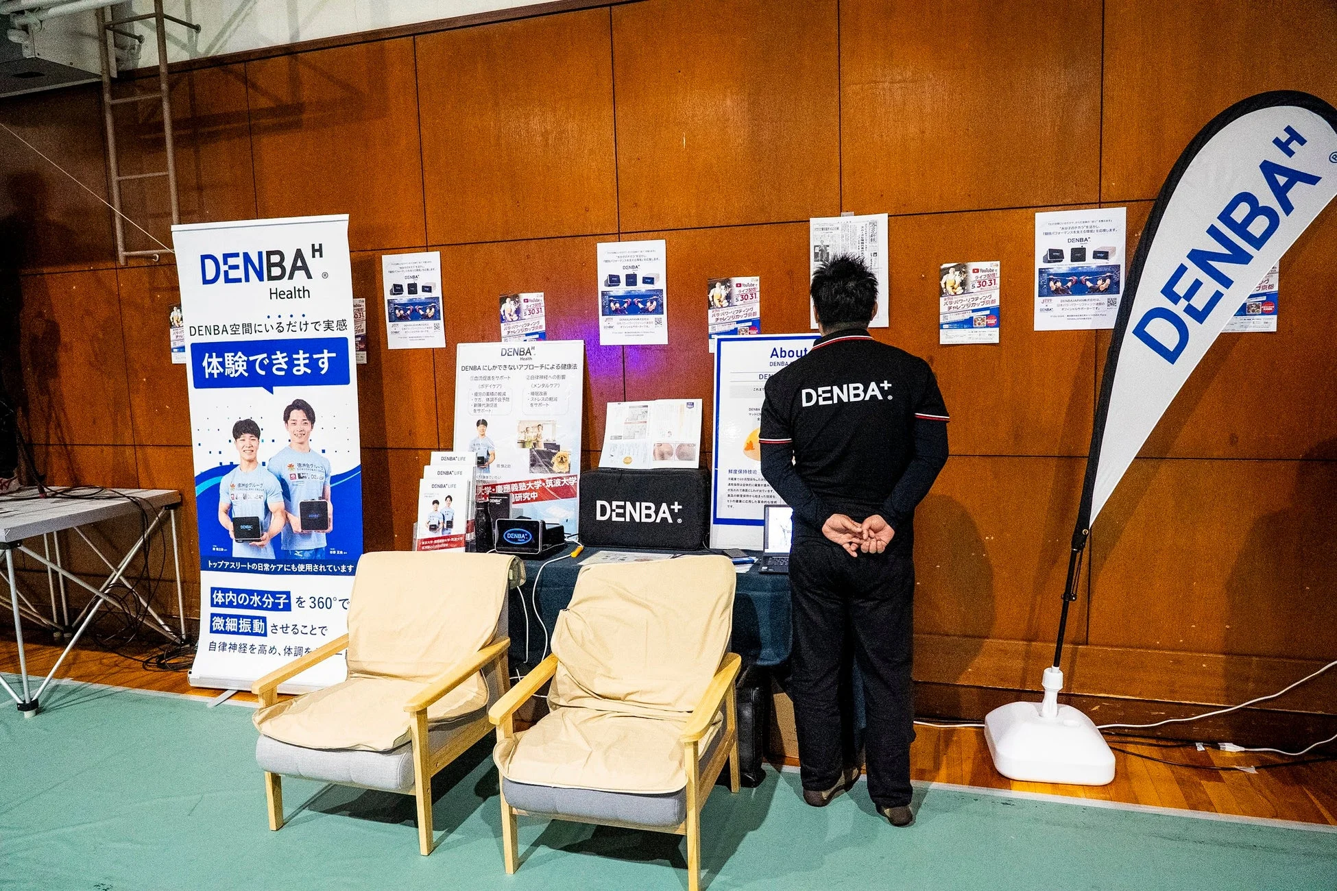 DENBA製品の展示ブースの様子。DENBA HealthとDENBA+という製品が紹介されており、体験可能なことや、血流促進、疲労回復、自律神経への影響など健康効果が説明されている。慶應義塾大学や筑波大学との共同研究も進行中であることが示されており、製品の信頼性をアピールしている。男性がブースに立っている。