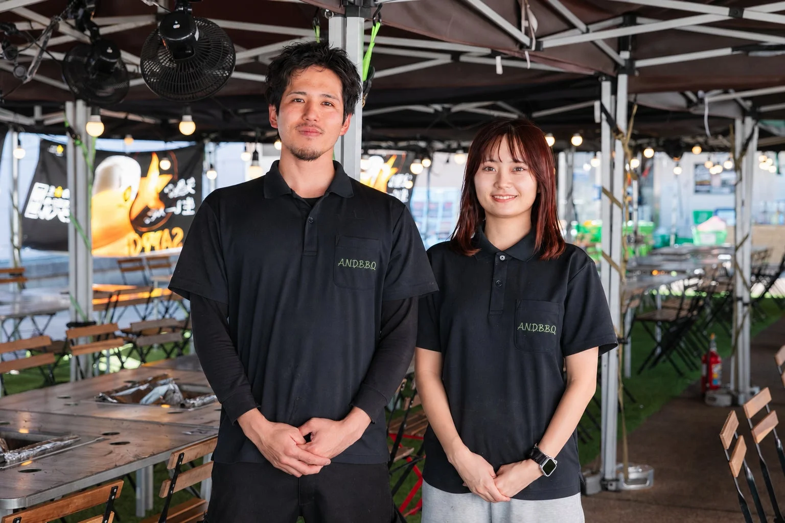 ANDBBQのロゴ入りポロシャツを着た男女スタッフが、屋外のバーベキューテラスで笑顔で立っています。背景には炭火焼肉を宣伝するバナーやテーブル席が見えます。