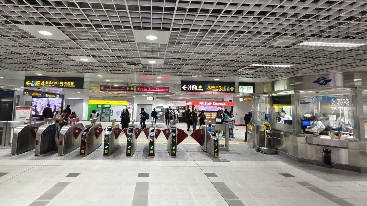 台北の地下鉄駅構内の光景で、複数の自動改札機が並び、多くの利用者が行き交っている。案内表示板やドーナツ店、駅員窓口も見られる。