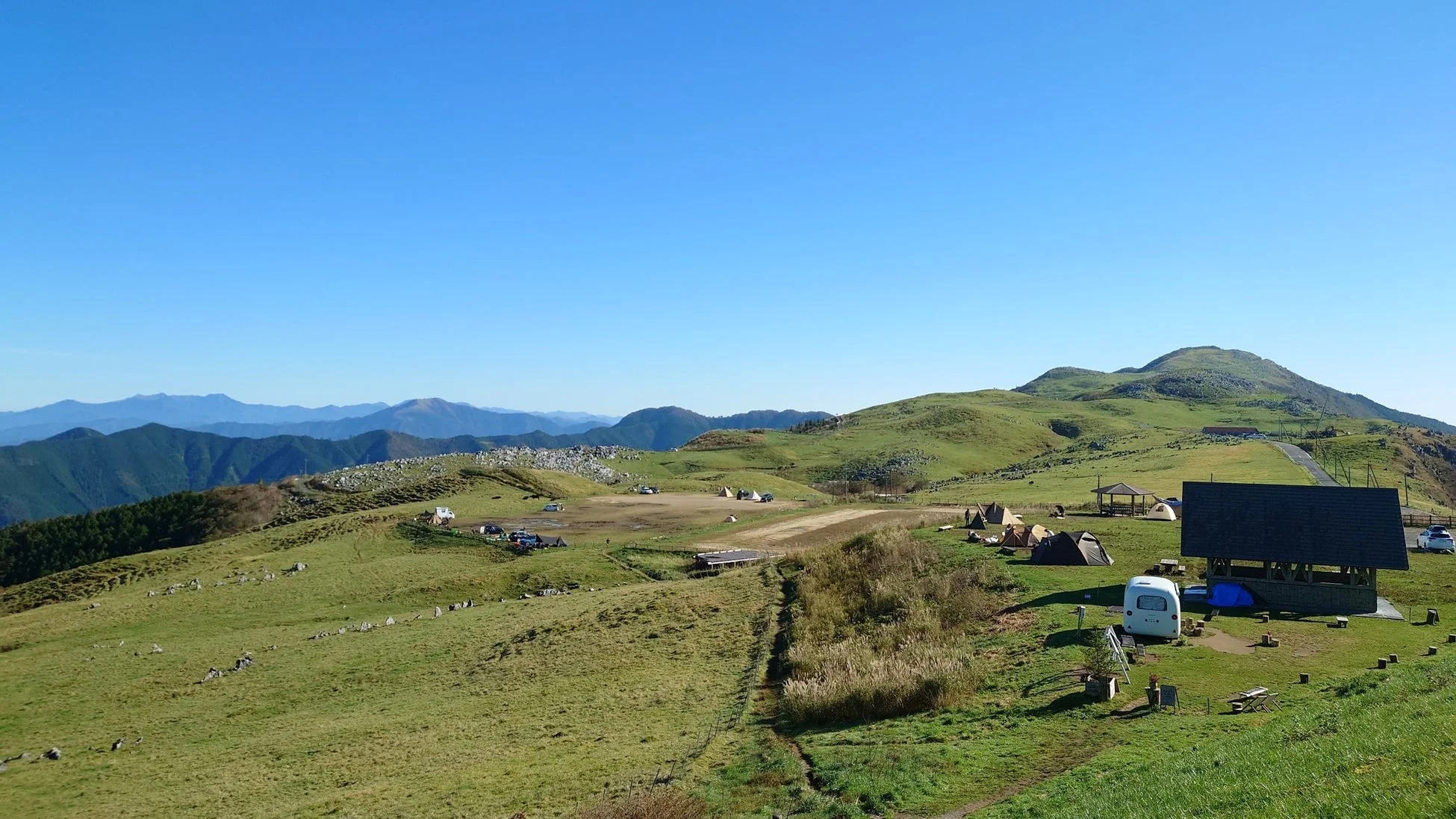 広大な緑の高原に広がるキャンプ場の風景。遠くには連なる山々が見え、テントや車が点在しています。