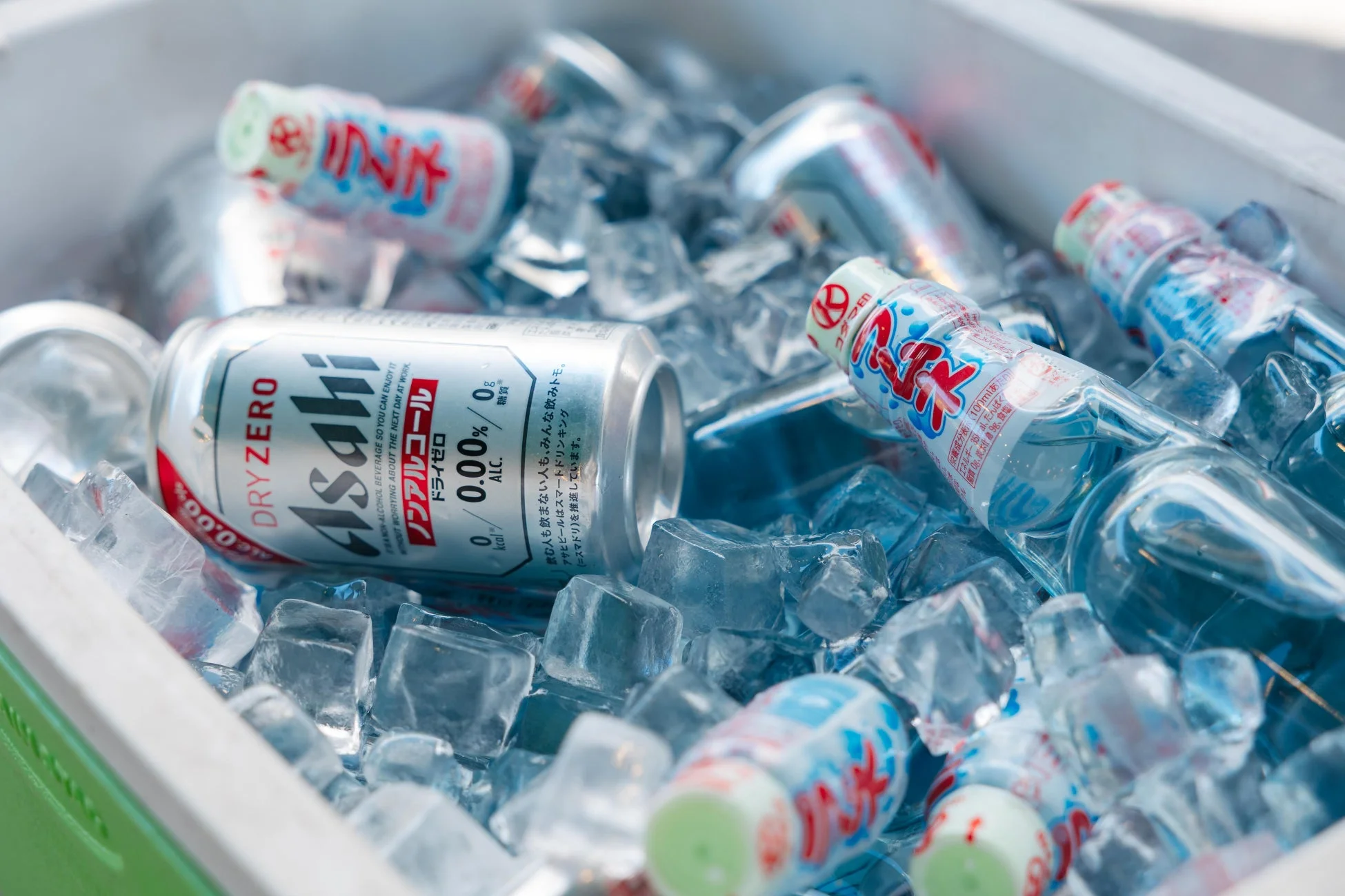 クーラーボックスにたっぷりの氷が敷き詰められ、アサヒドライゼロのノンアルコールビール缶と数本のラムネ瓶が冷やされている様子を捉えた画像です。夏のアウトドアやレジャーにぴったりの、涼しげな飲み物の光景が広がっています。