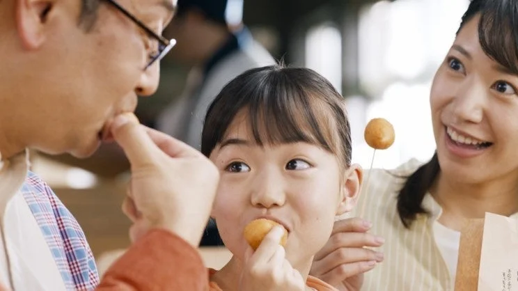 家族3人が集まって、笑顔で小さな丸いスナックを食べる楽しそうな一場面です。