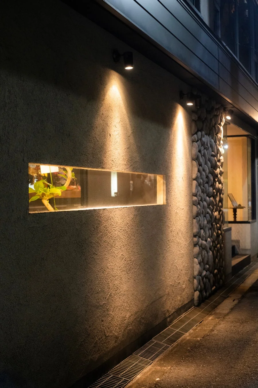 夜の建物の外壁が暖かなスポットライトで照らされています。壁には横長のスリット窓があり、中に植物が見えます。一部は石積みで装飾され、モダンで落ち着いた雰囲気を醸し出しています。