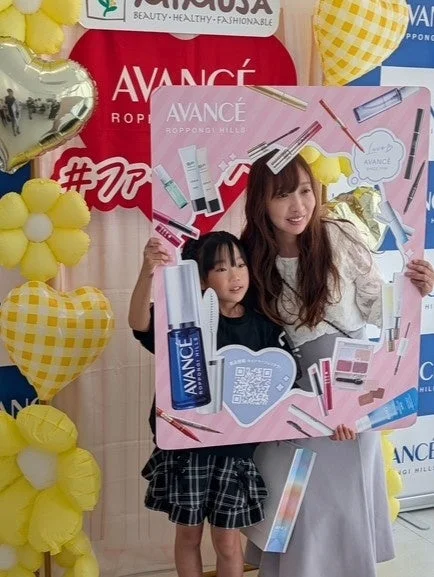 「AVANCÉ」のプロモーションイベントで、女性と子供が化粧品がデザインされたフォトフレームを持ち、笑顔でポーズをとっています。背景にはロゴやバルーンがあり、華やかな雰囲気です。