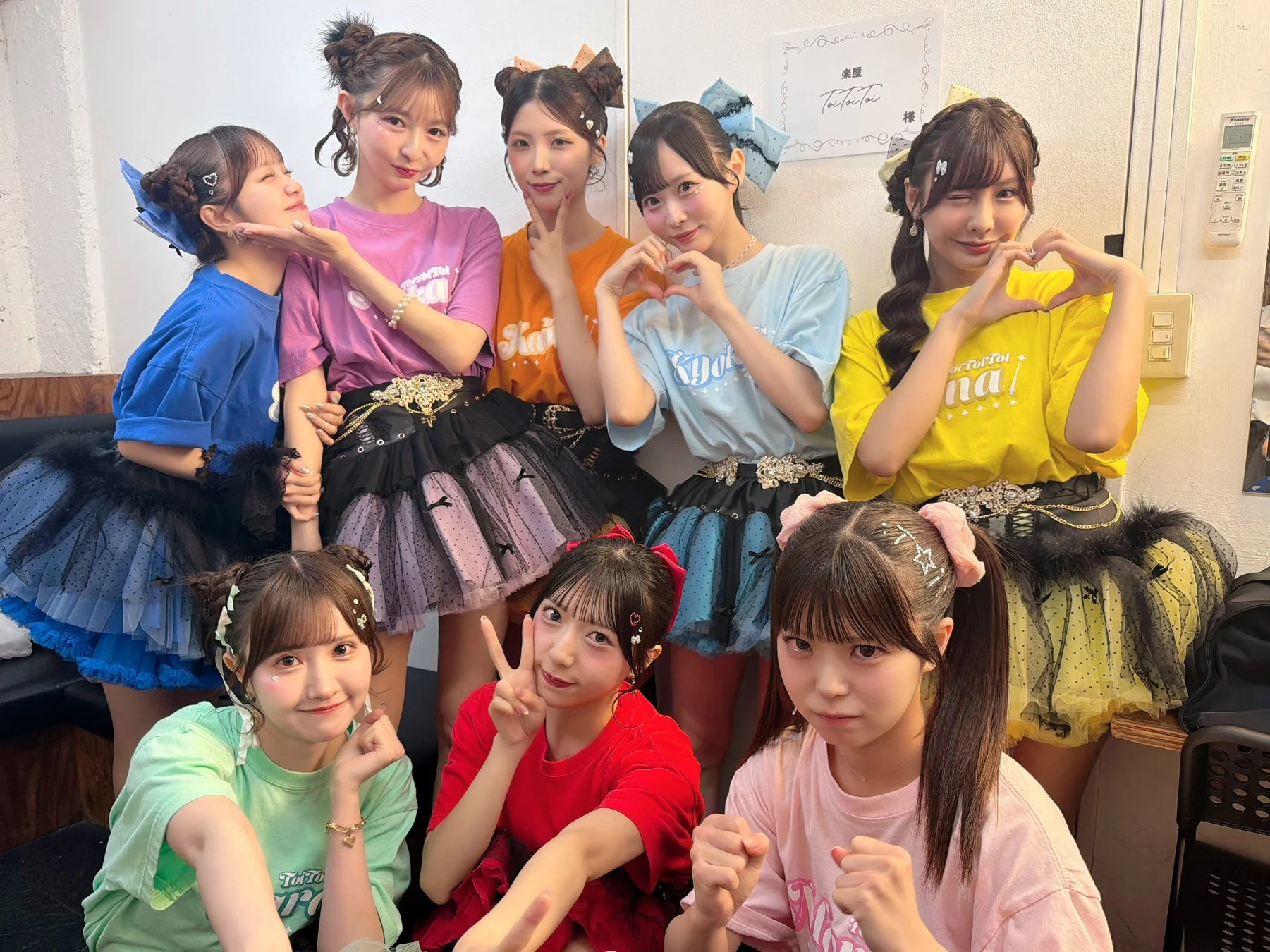 カラフルな衣装を身につけた9人の若い女性アイドルグループが楽屋で集合写真を撮っている。皆が笑顔でハートやピースサインなどのポーズをしており、和気あいあいとした雰囲気が伝わる。