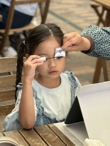 木製のテーブルに座った幼い女の子が、大人の手助けを受けながら眉毛をメイクしている様子。タブレットが横に置かれ、美容体験をしているようです。ツインテールの髪型が可愛らしい。