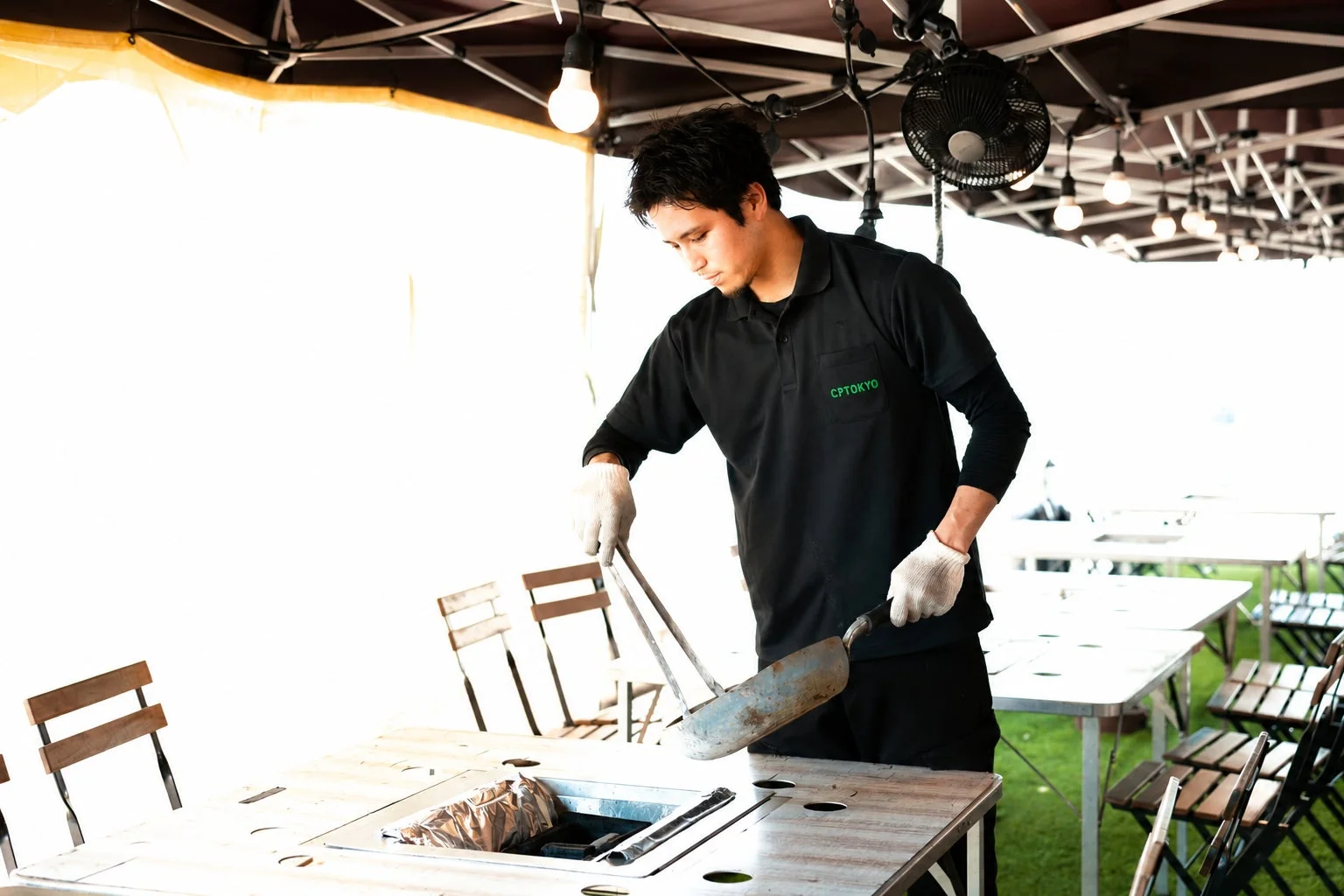 屋外のBBQ施設で、男性スタッフがグリルを準備している様子です。黒いポロシャツにグローブを着用し、道具を使って作業しています。
