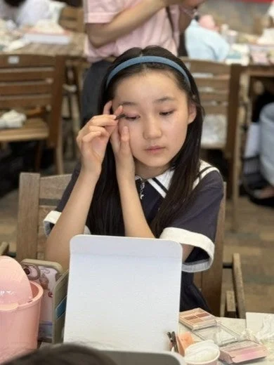若い女の子が鏡を見ながら、つけまつげかアイメイクをしている様子です。真剣な表情で、身だしなみを整えているように見えます。背景には他の人がいる室内空間です。