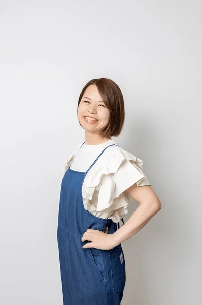 白い背景の前で、白いフリル袖のトップスとデニムのサロペットを着用した笑顔の日本人女性
