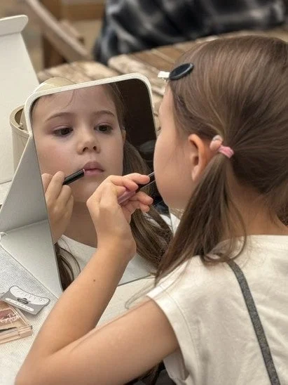 鏡を見ながら口紅を塗る小さな女の子の姿。おしゃれに興味を持つ子供の可愛らしい一瞬を捉えています。