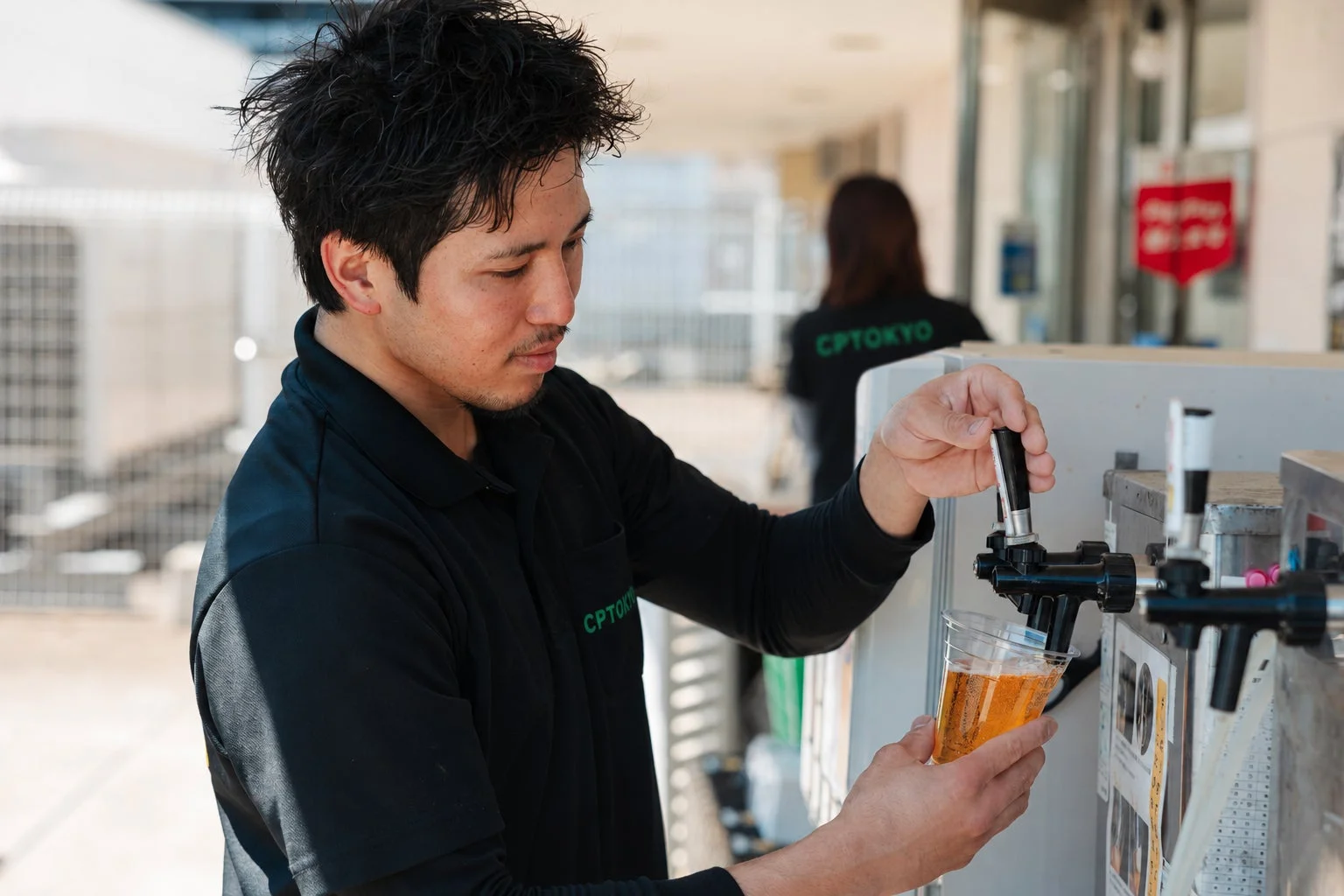 黒いポロシャツを着た男性がビールサーバーからプラスチックカップにビールを注いでいます。屋外のイベント会場のような場所で、手際よくドリンクを提供している様子です。