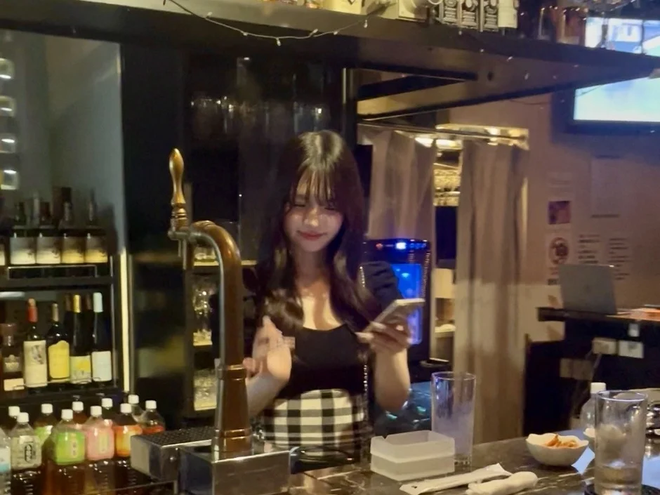 バーカウンターに立つ若い女性がスマートフォンを見て笑顔を見せています。店内にはビールサーバーや様々なお酒のボトルが並び、グラスやスナックが置かれ、夜のバーの活気ある雰囲気が感じられます。