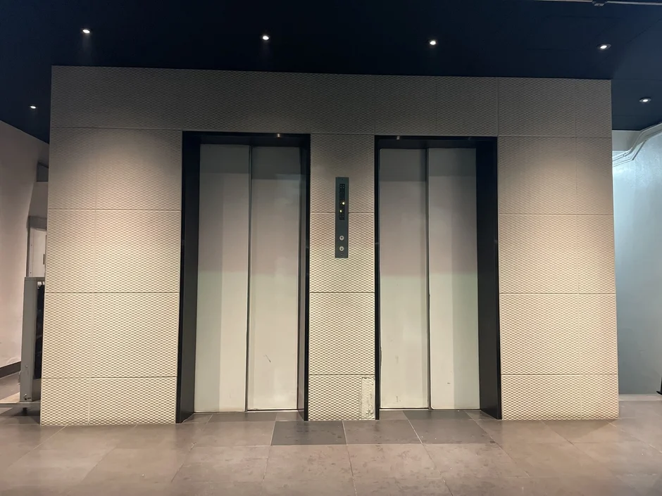 現代的な建物の内部にある、閉じた状態の2つのエレベーターのドアが写っています。壁は格子状のテクスチャで、天井には複数のダウンライトが見えます。