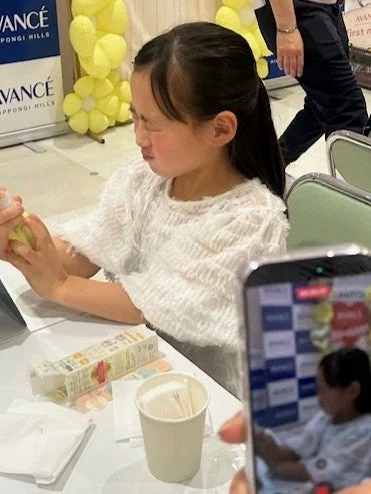 イベント会場らしき場所で、幼い少女が何かを試しながら顔をしかめている様子を捉えた一枚。手元には黄色いボトルがあり、スマートフォンでその様子が撮影されています。背景には「AVANCÉ ROPPONGI HILLS」の看板と黄色の風船が見えます。