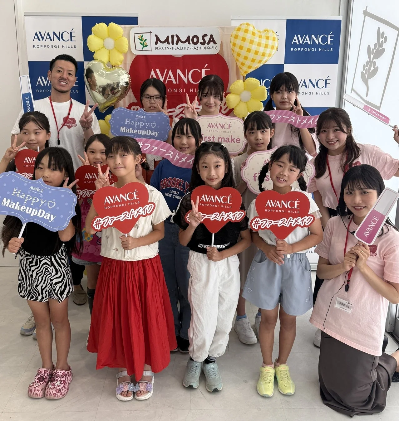 アヴァンセ六本木ヒルズ主催の「Happy MakeupDay」イベントで、子供たちが初めてのメイク体験を楽しむ集合写真です。参加者たちは笑顔で「#ファーストメイク」と書かれたプラカードを持ち、活気あるワークショップの様子が伝わります。